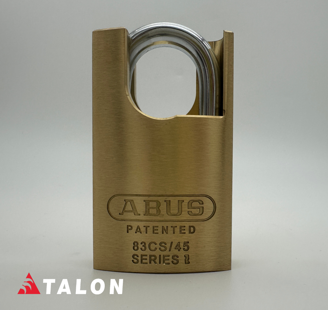 A brass padlock with a brushed metallic finish and a silver-colored shackle, featuring the brand name 'ABUS' and model '83CS/45 SERIES II' engraved on the front. A red and white 'TALON' logo watermark is visible in the bottom-left corner, set against a neutral gray background.